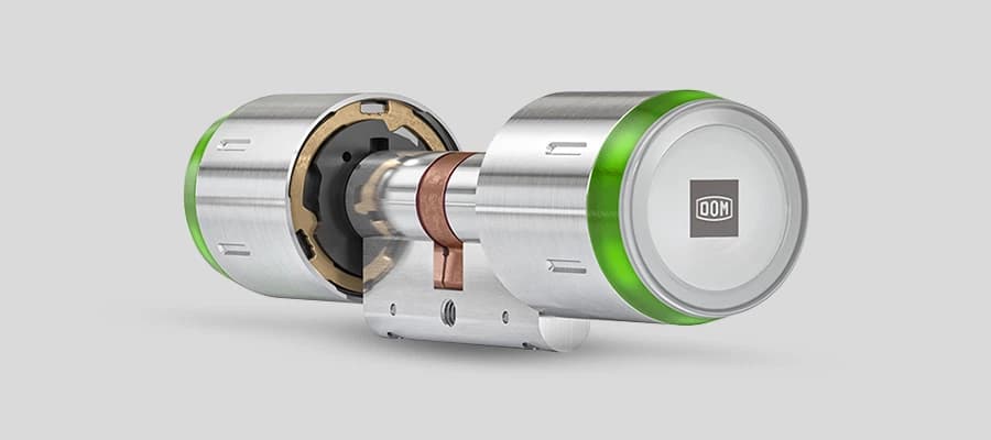 Moderner metallischer Türzylinder mit grünem LED-Ring und DOM-Logo auf grauem Hintergrund.