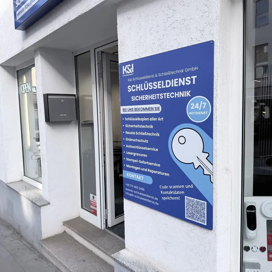 Eingang mit offenem Glasdoor und blauem Schild von K&I Schlüsseldienst mit Leistungsliste und QR-Code.